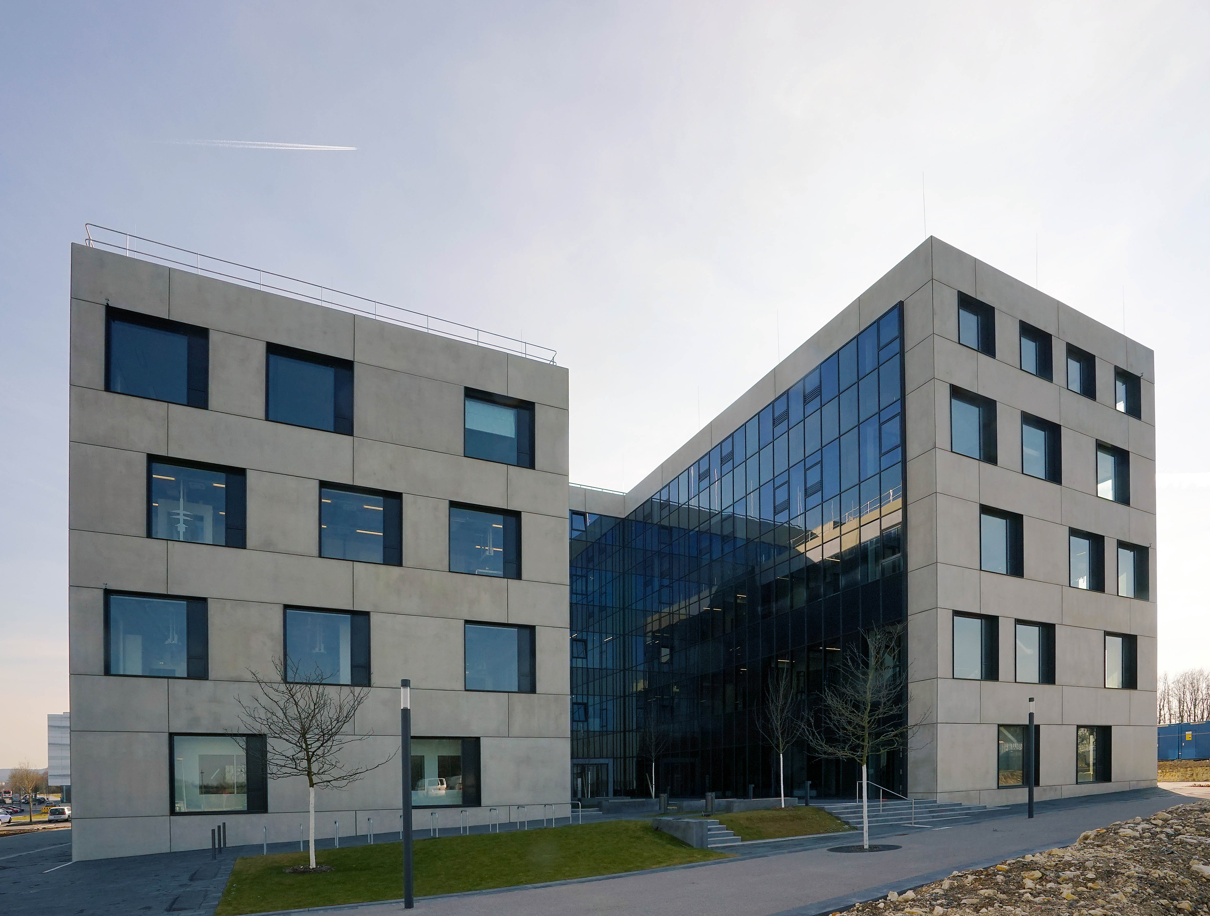 Leicht von Links betrachtet sieht man das U-förmige Gebäude hinter einer kleinen Grünfläche mit einem Treppenaufgang. Die Fassaden Rechts und Links sind sandsteinfarben gehalten und haben große rechteckige Fenster. Die Innenseite der rechten Gebäudehälfte zeigt eine gläserne Fensterwand.