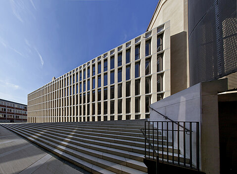 Philosophikum Münster Seitliche Ansicht des Philosophikums. Eine Treppe führt zum Gebäude. Die Fassade bildet die seitliche Gebäudeseite und besteht aus vielen Fenstern.