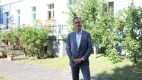 Männlich gelesene Person vor einer Fassade und Grünpflanzen.