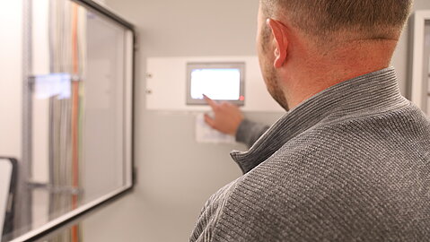 Person von hinten zeigt mit dem Finger auf ein kleines Display an einem technischen Schaltschrank.