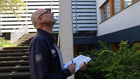 Mann mit Brille steht mit einem Klemmbrett vor einem mehrstöckigen Gebäude und blickt an der Fassade hinauf.