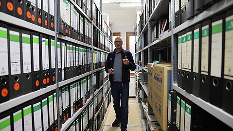 Mann steht zwischen zwei dicht befüllten Regalreihen mit schwarzen und grünen Aktenordnern in einem Archivraum.