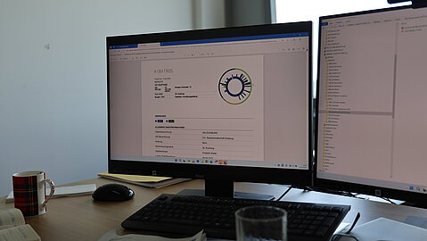 Büroarbeitsplatz mit zwei Monitoren – auf dem linken Bildschirm ist ein AiBATROS-Bericht zu sehen.