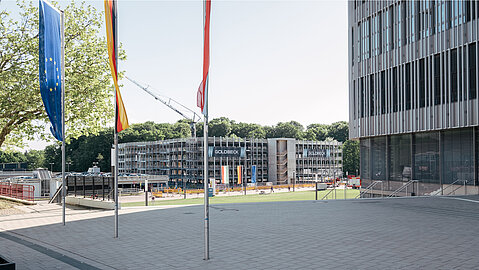 Blick vom Haupteingang des Universitätshauptgebäudes (UHG) der Universität Bielefeld auf die Baustelle des neuen Parkhauses – der Rohbau ist weit fortgeschritten, sichtbar mit „Goldbeck“-Kennzeichnung. Im Vordergrund wehen EU-, Deutschland- und Landesfahne.