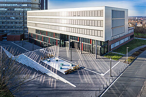 Ein Luftbild zeigt das Gebäude Q, eins von zwei neuen Universitätsbauten in Paderborn. Die Fassade besteht aus vielen Fenstern, die bei Sonnenstrahlen Licht auf den Vorplatz werfen. Da das Gebäude am Hang liegt, sieht man einen Treppenaufgang der vor einem großen Platz vor dem Haupteingang endet. Hier gibt es eine Fläche mit Sitzgelegenheiten. 