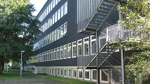 Seitliche Gebäudeansicht der ehemaligen pädagogischen Hochschule in Essen. Man sieht eine Außentreppe sowie eine graue Fassade mit vielen Fenstern.