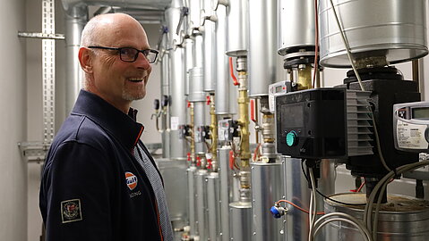 Mann mit Brille steht lächelnd vor einer technischen Anlage mit Rohren, Ventilen und einer Pumpensteuerung.