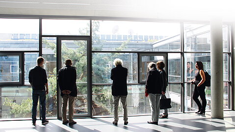 Fünf Personen stehen an einer Fensterfront im Gebäude und schauen in einen begrünten Innenhof hinunter.