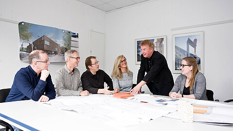Die sechs-köpfige Projektgruppe sitzt um einen weißen Tisch herum und bespricht zusammen mit dem Projektmanager die vorliegenden Pläne. Die Personen sind der Projektleitung zugewandt. Der Projektmanager deutet auf einen Plan und ist einer Person zugewandt.