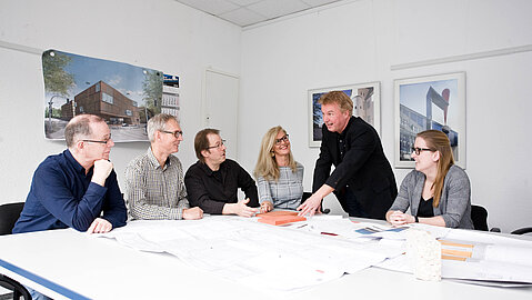 Die sechs-köpfige Projektgruppe sitzt um einen weißen Tisch herum und bespricht zusammen mit dem Projektmanager die vorliegenden Pläne. Die Personen sind der Projektleitung zugewandt. Der Projektmanager deutet auf einen Plan und ist einer Person zugewandt.