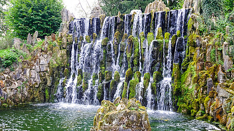 Ein künstlich angelegter Wasserfall mit moosbewachsenen Steinen und Wasserbecken davor. 