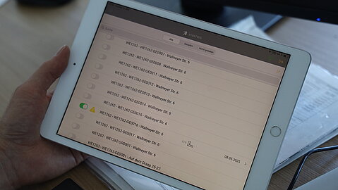 Hand hält ein Tablet mit der AiBATROS-App, auf der eine Liste von Gebäudeobjekten erkennbar ist. 