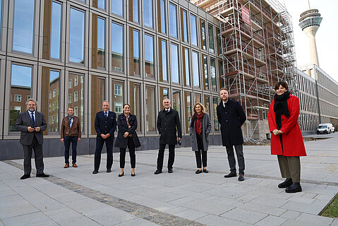Eine Gruppe von beteiligten Akteuren ist vor dem Polizeipräsidium Düsseldorf zusammengekommen und freut sich über die Einweihung des ersten Bauabschnitts. Im Hintergrund sieht man den fertiggestellten Neubauteil und den eingerüsteten Altbau, der im zweiten Bauabschnitt fertiggestellt wird.