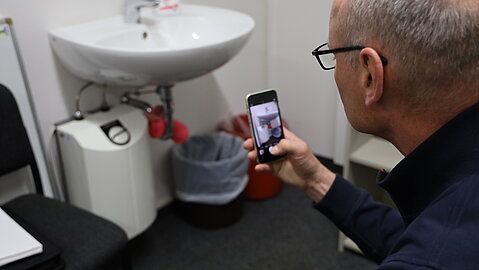 Mann fotografiert mit dem Smartphone die Unterseite eines Waschbeckens mit sichtbaren Leitungen.