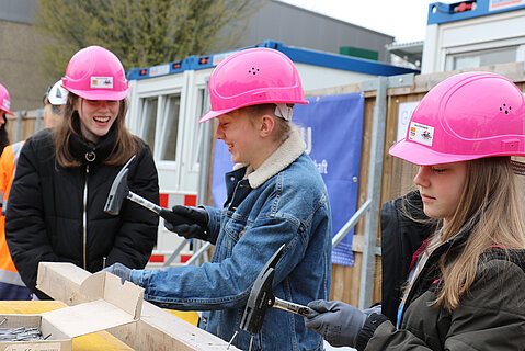 Drei Mädchen mit einem pinken Bauhelm schlagen mit einem Hammer Nägel ins Holz. Das Bild wurde auf einer Baustelle aufgenommen.