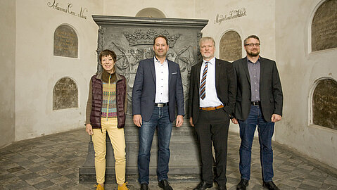 Vier Personen stehen in der Fürstengruft im Unteren Schloss Siegen vor einem denkmal. An den Wänden sind einzelne Tafeln zu erkennen.