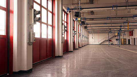 In der Halle für die Einsatzfahrzeuge befinden sich neben jedem der Ausgangstore Ampelanlagen. Die Türen haben kleine quadratische Fenster. Am Ende der leeren Halle befindet sich ein Treppenaufgang. 