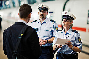 Eine Polizeibeamtin und ein Polizeibeamter befragen einen Passanten. Die Beamten sind der Kamera zugewandt. Die Beamtin deutet auf ein Dokument in ihrer Hand. Im Hintergrund befindet sich ein ICE der Deutschen Bahn. 