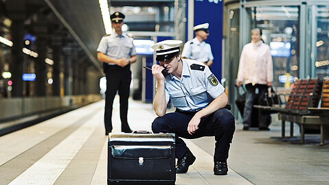 Ein uniformierter Polizist hockt vor einem Koffer und spricht durch ein Walkie Talkie. Im Hintergrund kontrolliert ein weiterer Beamte die Situation und ein dritter Polizist ist einer Passantin zugewandt.