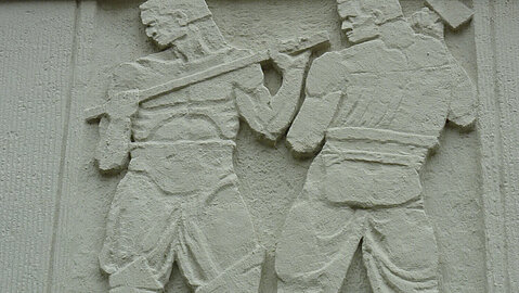 An der Fassade des Gebäudes werden Berufe aus dem Steinbruch dargestellt. Hier sind zwei Bergarbeiter bei ihrer Arbeit zu sehen.