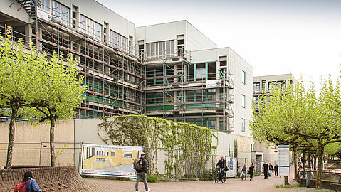 Der Bau wird von der Seite aus betrachtet. Hinter einer begrünten Mauerfassade liegt das neuen Universitätsgebäude. Am Gebäude befindet sich ein Baugerüst. Auf einem bepflanzten Innenhof, der parallel zum Universitätsgebäude verläuft befinden sich mehrere Personen.