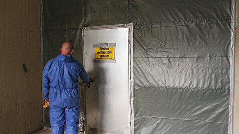 Eine in Schutzanzug gekleidete Person betritt einen abgesprerrten Gebäudebereich, der mit einem Warnschild gekennzeichnet ist. 