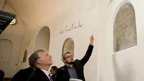 Eine Person deutet mit einem Arm auf eine Inschrift oberhalb eine in der Wand integrierten Tafel, während eine andere Personen nach oben blickt. Im Hintergrund sind weitere Personen zu erkennen.