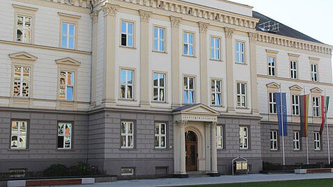 Ministerium der Justiz NRW Das Ministerium der Justiz NRW ist im Klassizismus entstanden. Im unteren Drittel des Hauses sind Fenster in eine graue Fassade eingelassen. Die darüber befindenden Etagen sind im hellen Stein gehalten. Es gibt Stuckelemente und der Eingangsbereich hat zu beiden Seite Säulen. Außerdem zu sehen sind Fahnen sowie Rasen vor dem Gebäude.