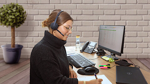 Eine Mitarbeiterin sitzt an einem Schreibtisch vor ihrem Computer. Sie ist im Home Office und telefoniert über ihr Headset.