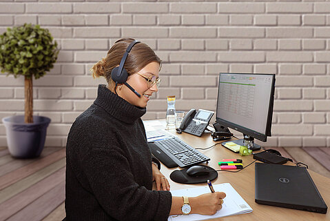 Eine Mitarbeiterin sitzt an einem Schreibtisch vor ihrem Computer. Sie ist im Home Office und telefoniert über ihr Headset.