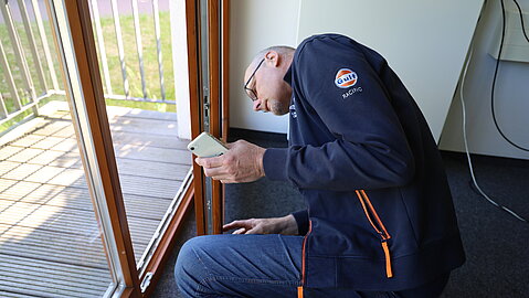 Mann kniet auf dem Boden und fotografiert mit seinem Smartphone den unteren Bereich eines Holzfensters mit Balkonzugang.