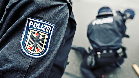 Im Bildvordergrund ist das Armabzeichen eines Bundespolizisten zu sehen. Das Abzeichen zeigt den Schriftzug und das Adlersymbol der Polizei. Dahinter ist ein uniformierter Polizist mit Helm erkennbar, der eine Festnahme am Boden durchführt.