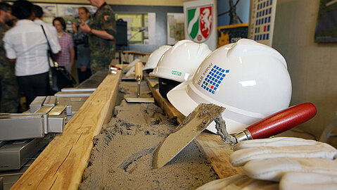 In einer Reihe liegen weiße Baustellenhelme mit dem Logo des BLB NRW auf einem Holzbalken. Davor ist Beton zu sehen, auf dem eine Kelle liegt. Im Hintergrund sind mehrere Personen erkennbar.