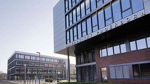 Die beiden neuen Gebäudeteile I und Q der Paderborner Universität bilden zusammen den Eingang zur Uni. Auf dem rechten Gebäude im Bild ist der Schriftzug der Universität in großen Lettern angebracht. Ein Metallbau mit langen horizontalen Fensterreihen überlagert den unteren Gebäudeteil und bildet einen kurzen Vorsprung. Vor dem Gebäude liegt eine Grünfläche