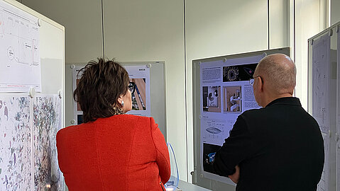 Eine Person in einer roten Jacke und eine Person in einem dunklen Pullover stehen vor einer Infowand.