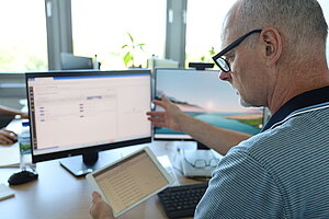 Mann sitzt mit einem Tablet in der Hand am Schreibtisch, während er auf die Software-Oberfläche seines Monitors zeigt.