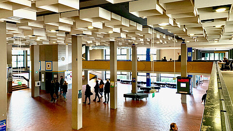 Einblick in die technische Hochschule Ostwestfalen-Lippe am Standort Lemgo. An der Decke sind viele Kästchen eingelassen in denen zum Teil Lampen integriert sind. Es sind Säulen zu sehen, zwischen den viel Platz ist sowie mehrere Sitzmöglichkeiten.