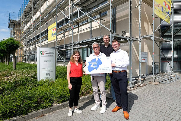 Vier Personen präsentieren vor dem Finanzamt das neue Hinweisschild zur nachhaltigen Energieversorgung.