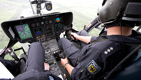 Im Cockpit sitzen zwei uniformierte Beamte der Bundespolizei. Zwischen ihnen befindet sich die Steuerkonsole mit Flugelektronik. Ein kleiner Kompass zeigt die Himmelsrichtung an. Die Piloten fliegen über ein Waldareal.