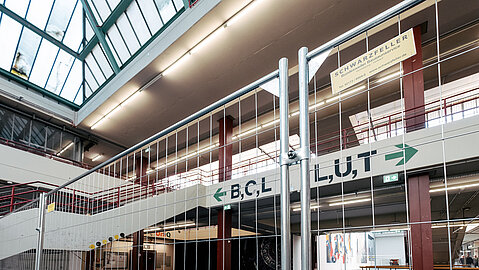 Baustelleneinrichtung im Universitäts-Hauptgebäude (UHG) Bielefeld während des 2. Bauabschnitts. Eine Absperrung trennt den Bereich ab, Alte Wegweiser für die Gebäudeteile B, C, L sowie L, U, T sind sichtbar. Die Sanierungsarbeiten finden im laufenden Betrieb statt.