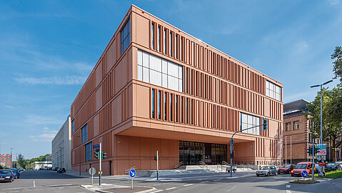 An einer Straßenkreuzung liegt das orangene Gebäude des Justizzentrums in Bochum. Der Eingang ist unter einem Überbau über einen Treppenaufgang ereichbar. Die Fensterfronten sind sowohl großflächig, als auch schmal vertikal nebeneinander verordnet. Das gebäude ist so konstruiert, dass der Eingangsbereich überdacht ist.