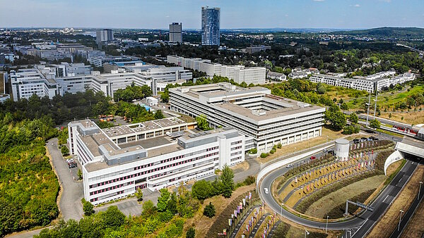 Luftbild eines aus mehreren Teilen bestehenden Gebäudes mit heller Fassade. Im Hintergrund weitere Gebäude.