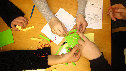 Beim Girls Day sollte etwas gebastelt werden. Man sieht hier mehrere Hände, die Papier und Stäbchen halten. Weiteres Bastelmaterial liegt auf dem Tisch bereit.