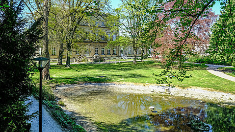 Im Vordergrund liegt ein kleiner Teich, während sich im hinteren Teil, versteckt hinter Bäumen das Palaisgebäude zeigt.
