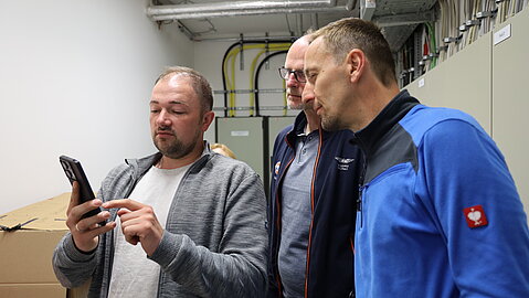 Drei Männer stehen in einem Technikraum mit Kabeltrassen und schauen gemeinsam auf ein Smartphone.