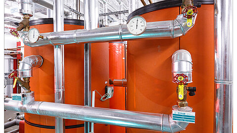 Mehrere Rohre, Leitungen und Ventile führen durch orangene fassförmige Behälter. Die Rohre verlaufen an der Decke weiter.