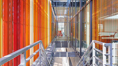 In der Mitte des Bildes führt eine Treppen mit einem Geländer rechts und links hinunter in einen Raum. Rechts und links sind dekorative Elemente in Orange- und Brauntönen wie senkrechte Lamellen angeordnet.